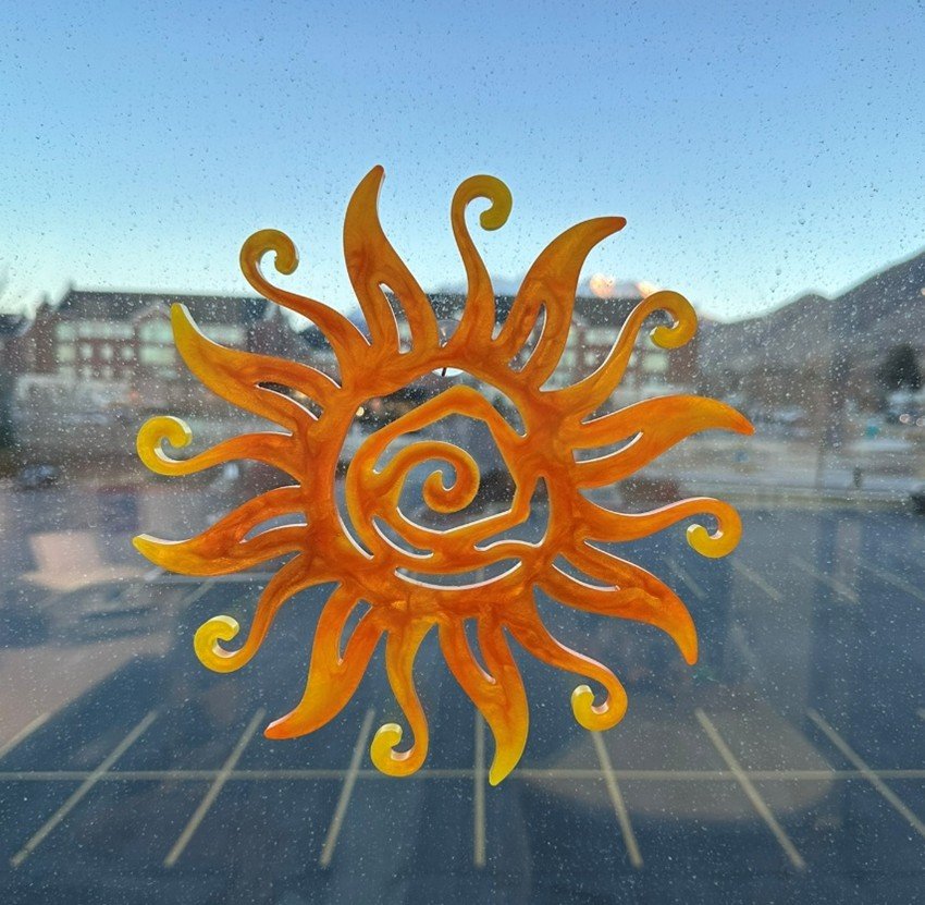 A stylized sun-shaped glass art piece hanging in a window with blue and golden tones